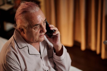 Téléchargez les photos : Portrait of senior man in pajamas sitting on bed and taking on phone - en image libre de droit