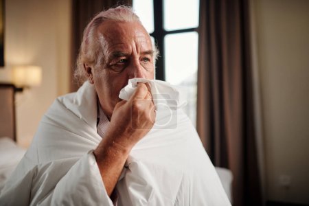 Foto de Portrait of senior man wrapping in duvet and blowing nose - Imagen libre de derechos