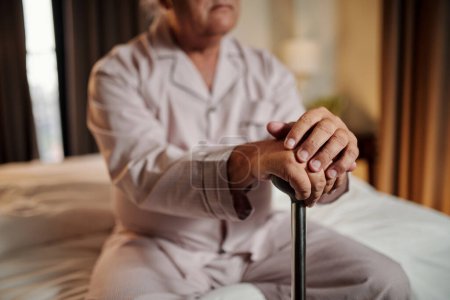 Téléchargez les photos : Homme âgé en pyjama assis sur le lit avec un bâton de marche - en image libre de droit