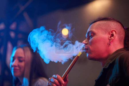 Foto de Elegante joven fumando narguile cuando pasa la noche en el club nocturno con amigos - Imagen libre de derechos