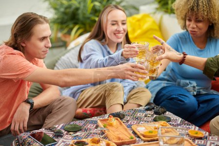 Photo for Group of young people toasting with glasses of beer over table with snacks - Royalty Free Image