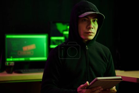 Foto de Serious hacker in cap and black hoodie holding tablet computer - Imagen libre de derechos