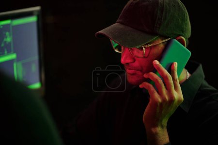 Téléchargez les photos : Hacker in cap and glasses talking on phone, when working in dark room with neon light - en image libre de droit