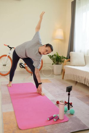 Téléchargez les photos : Femme suivant tutoriel sur smartphone lors de la pratique du yoga à la maison - en image libre de droit