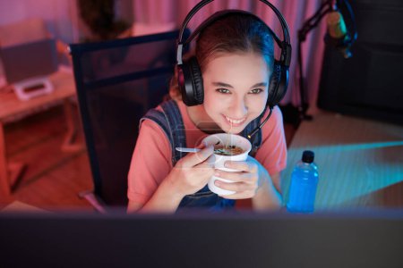 Foto de Smiling teenage girl eating ramen when watching lets play video on computer late at night - Imagen libre de derechos