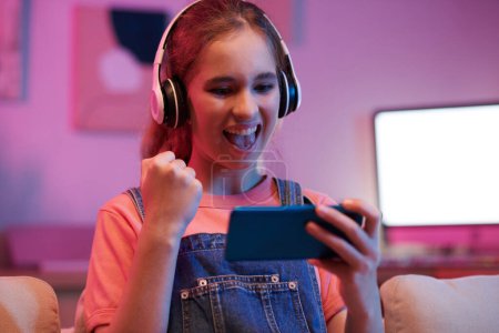 Téléchargez les photos : Adolescente excitée faisant geste de pompe poing lors de la célébration jeu gagnant sur smartphone - en image libre de droit