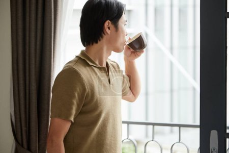 Foto de Pensativo joven bebiendo taza de café de la mañana y mirando a través de la ventana - Imagen libre de derechos