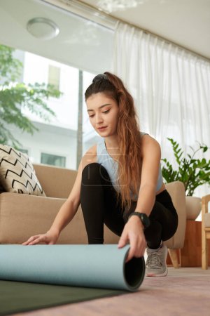 Foto de Young woman rolling out yoga mat on floor in living room - Imagen libre de derechos