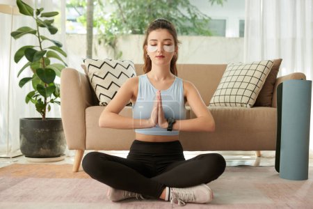Téléchargez les photos : Jeune femme assise en position lotus, en gardant les mains en geste namaste lors de la méditation à la maison - en image libre de droit