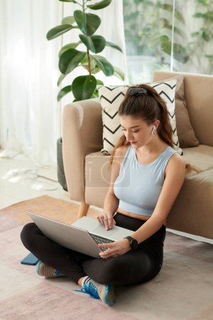 Téléchargez les photos : Young woman sitting on floor in living room and searching for online fitness class - en image libre de droit