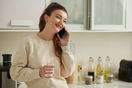 Foto de Portrait of young smiling woman drinking glass of fresh water and talking on phone - Imagen libre de derechos