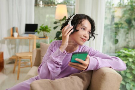 Téléchargez les photos : Adolescente positive reposant sur un canapé à la maison, écoutant de la musique et regardant par la fenêtre - en image libre de droit