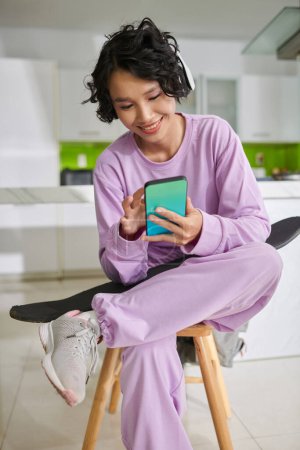 Foto de Smiling teenage girl sittingo on stool and answering text messages on smartphone - Imagen libre de derechos
