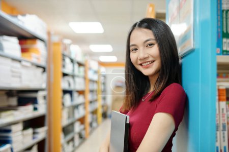 Téléchargez les photos : Portrait of smiling high school student with laptop standing in library - en image libre de droit