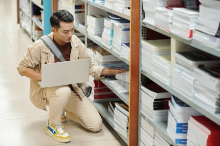 Téléchargez les photos : High school student with laptop searching for books for his research in library - en image libre de droit