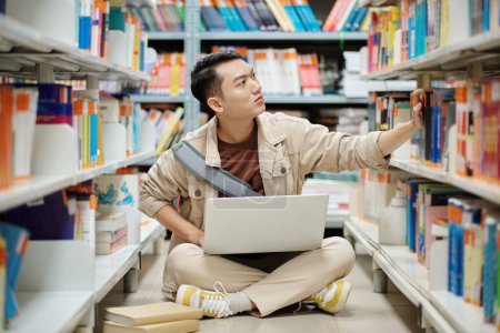 Photo for School student sitting on library floor and taking book from shelf - Royalty Free Image