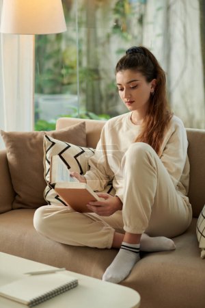 Téléchargez les photos : Jeune femme en vêtements de salon assis sur le canapé et le livre de lecture pour la classe à venir - en image libre de droit