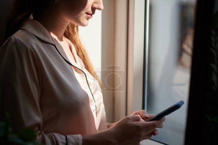 Téléchargez les photos : Souriante fille debout à la fenêtre et SMS ami - en image libre de droit
