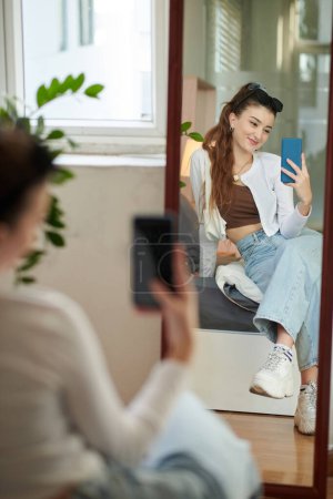 Téléchargez les photos : Smiling girl sitting in front of mirror and taking selfie - en image libre de droit
