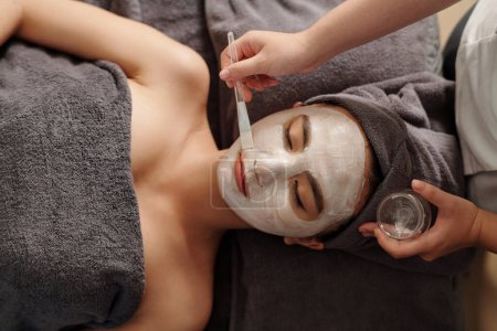 Foto de Cosmetologist applying brightening white clay mask on face of young woman - Imagen libre de derechos