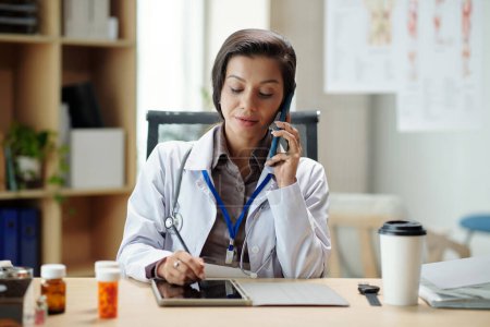Photo for General practitioner talking on phone with client and taking notes in form on digitial tabelt - Royalty Free Image