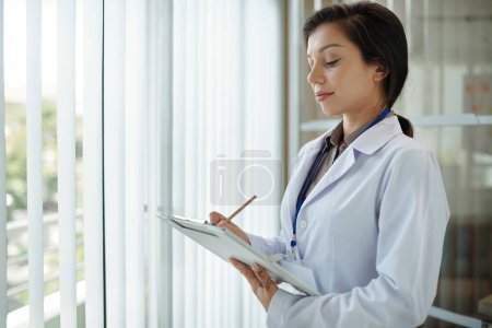 Foto de Médico general sonriente parado en la ventana de la oficina y llenando el documento en el portapapeles - Imagen libre de derechos