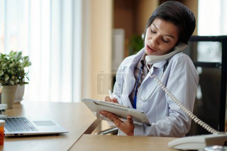 Photo for General practitioner talking on phone with patient and filling form on clipboard - Royalty Free Image