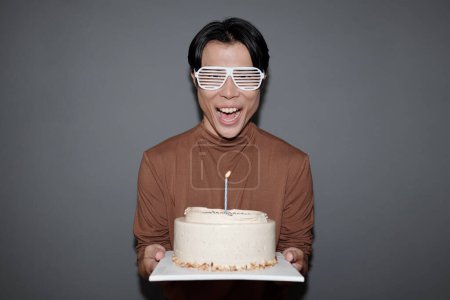 Photo for Excited young man in party glasses holding birthday cake with one candle - Royalty Free Image