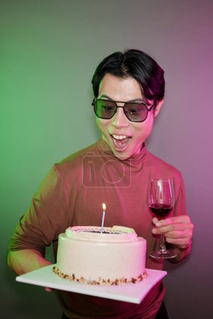 Téléchargez les photos : Émotionnel excité jeune asiatique homme avec verre de vin regardant gâteau d'anniversaire - en image libre de droit
