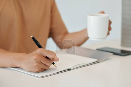Téléchargez les photos : Image rapprochée du jeune homme buvant une tasse de café le matin et écrivant des plans pour la journée - en image libre de droit