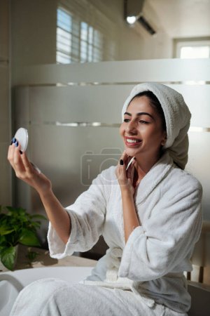 Téléchargez les photos : Sourire jeune femme regardant un miroir compact lors de l'application de lotion hydratante après avoir pris un bain - en image libre de droit