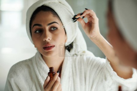 Téléchargez les photos : Jeune femme appliquant un sérum éclaircissant visage en face du miroir de salle de bain - en image libre de droit