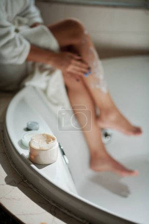 Foto de Tarro con rica mantequilla corporal nutritiva en el borde de la bañera, mujer aplicándolo en el fondo - Imagen libre de derechos