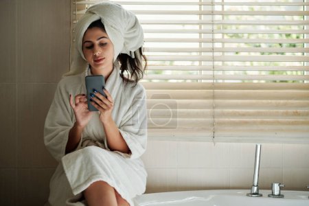 Téléchargez les photos : Jeune femme en peignoir vérifiant les médias sociaux après avoir pris un bain le soir - en image libre de droit