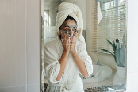 Téléchargez les photos : Jeune femme lavant masque d'argile et regardant miroir - en image libre de droit
