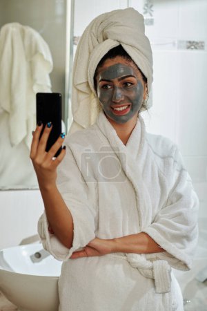 Foto de Mujer feliz fotografiándose con máscara de arcilla en la cara - Imagen libre de derechos