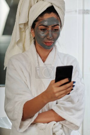 Téléchargez les photos : Jeune femme avec masque charbon de bois sur le visage vérification des médias sociaux sur smartphone - en image libre de droit