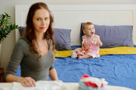 Téléchargez les photos : Joyeux babygirl assis sur le lit lorsque la mère repassage vêtements - en image libre de droit