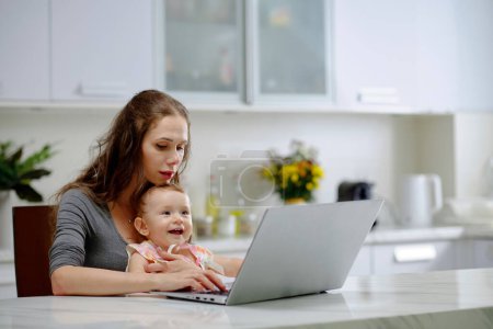 Photo for Young woman working on laptop with little daughter sitting on her laps - Royalty Free Image