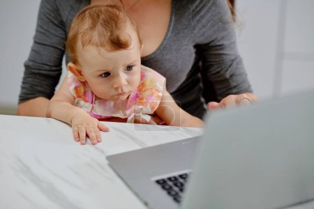 Téléchargez les photos : Curieux petite fille regardant l'écran d'ordinateur portable lorsque sa mère travaille à la maison - en image libre de droit