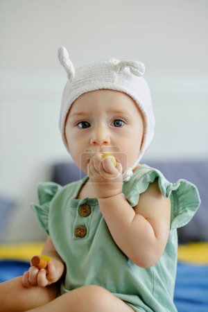 Photo for Cute little girl in funny hat sitting on bed and sucking wooden toy - Royalty Free Image