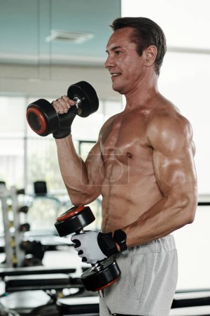 Foto de Sonriente hombre maduro en forma haciendo ejercicio bíceps con pesas - Imagen libre de derechos