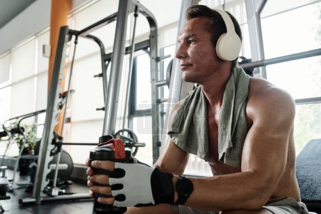 Photo for Fit mature man enjoying good music amd drinking water when resting after deadlift - Royalty Free Image