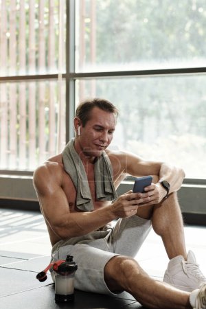 Photo for Sportsman sitting on gym floor and choosing music for training from playlist on smartphone - Royalty Free Image
