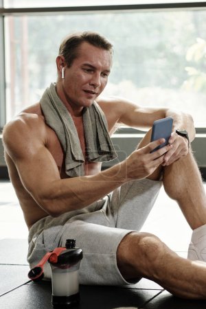 Téléchargez les photos : Fit homme assis sur le sol de la salle de gym et textos avec entraîneur personnel - en image libre de droit