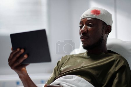 Foto de Soldado herido afroamericano con la cabeza herida viendo vídeo en la tableta PC mientras está acostado en el hospital - Imagen libre de derechos