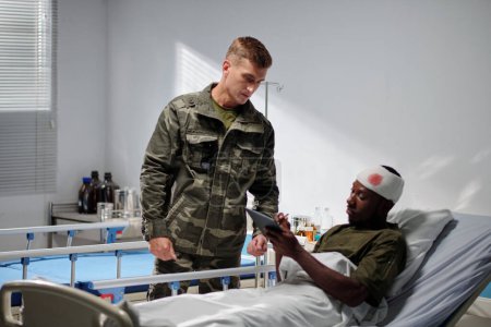Téléchargez les photos : Soldat en uniforme rendant visite à son ami blessé à l'hôpital, il lui montre des photos sur tablette numérique - en image libre de droit