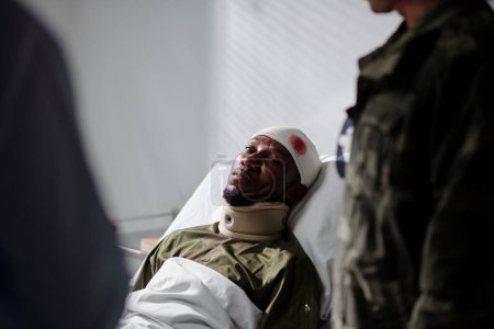Foto de Soldado herido afroamericano con la cabeza herida acostado en la cama en el hospital mientras el médico y su colega lo visitan - Imagen libre de derechos