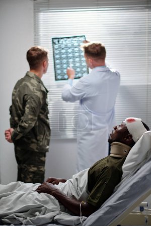 Foto de Médico profesional apuntando a la imagen de rayos X y discutiéndolo con un soldado con un hombre herido acostado en la cama en la sala - Imagen libre de derechos
