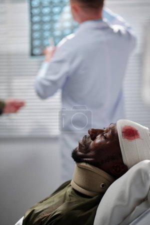 Photo for Wounded African American patient with injured head lying on bed in ward with doctor examining x-ray image in background - Royalty Free Image